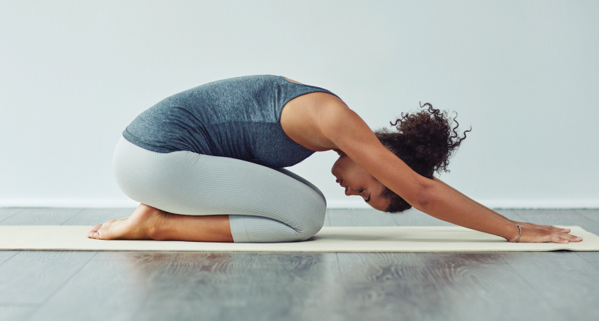 A woman sits in child's pose on a gym mat, stretching her lower back, arms extended.